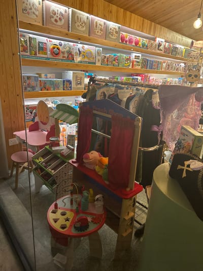 The image depicts an interior scene, likely a children's toy store, boutique, or play area, located in Egkomi Lefkosias, Cyprus. The space is illuminated by artificial light, suggesting an indoor setting during the day.

On the left, a wall lined with wooden shelving units showcases a wide array of packaged children's toys and educational products. These items include colorful textured balls, building blocks, and various activity sets, some bearing text such as "J'UDI," "ACTIVITE," and "ACTIVITY GAR" on their packaging.

The central and right sections of the image are dominated by an engaging children's play area. A prominent blue and red wooden structure, designed as a "Fresh Shop" stall or puppet theater with red curtains, contains several plush toys, including a notable pink pig. Beside it, a green and white striped umbrella signals another play zone, possibly a market stand or kitchen, furnished with child-sized pink wooden chairs and tables. A large, round, multi-colored activity table is also visible, featuring manipulative elements like gears, a bead maze, and shape sorters.

In the background, further displays of children's merchandise can be seen, including hanging garments with lace details and a distinctive black hat with gold embroidery. The overall impression is one of an organized, colorful, and inviting environment tailored for children's play and exploration. No individuals are visible in the scene.
