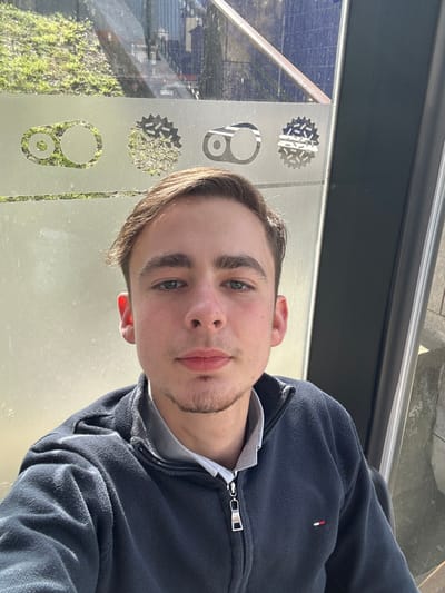 A portrait-oriented photograph features a young Caucasian male, approximately 18-25 years old, with short brown hair, blue-green eyes, and a light goatee beard, looking directly at the camera with a neutral expression. He is wearing a navy blue quarter-zip fleece jacket over a light grey collared shirt; a small red, white, and blue rectangular logo is present on the left chest of the fleece. The subject is situated next to a large glass window or partition adorned with frosted or etched circular motifs resembling bicycle components, specifically chain links and sprockets, consistent with the Crankbrothers brand. Through the glass, green foliage, urban structures, and reflections of sunlight are visible in the background. A dark vertical frame element is present on the right edge of the image. The scene is identifiable as being inside the Bike Bar, located in Parque Infante D. Pedro, Aveiro, Portugal.