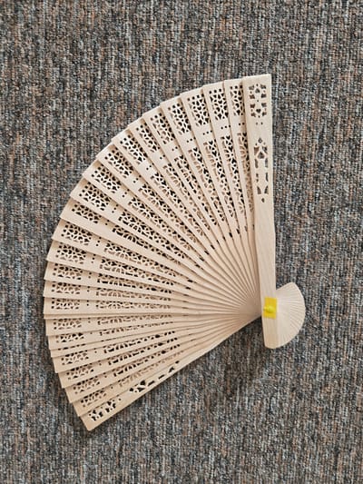 The media file displays a close-up, top-down view of a light-colored, wooden folding fan resting on a textured, greyish-brown carpet. The fan is partially opened, fanned out in a semi-circular shape. The wood is intricately carved with a decorative pattern of small, repeating geometric shapes and dots along the edges of each segment. The handle of the fan is visible at the top, closed and secured with a small, bright yellow tab or sticker. The fan is positioned diagonally across the frame, with the handle towards the upper right and the fanned-out portion extending towards the lower left. The lighting appears even, suggesting an indoor setting with diffused light, possibly during the daytime. There is no visible text in the image. The scene is static, with no activity taking place. The focus is solely on the object and its texture. The carpet background has a flecked appearance, with variations in shades of grey, brown, and hints of other muted colors.