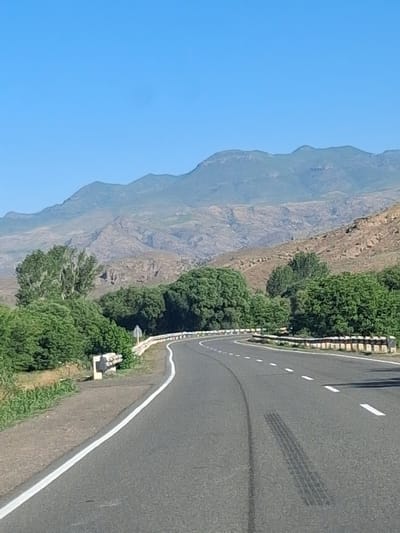 The image shows a road curving through a mountainous, arid landscape. The road is paved with asphalt and has white lane markings. There are guardrails along the side of the road, and vegetation, including trees and bushes, is visible on either side. In the background, rugged mountains rise under a clear blue sky. There are no people or vehicles visible in the scene. The scene appears to be a rural or remote location, possibly a mountain pass or highway. The presence of tire marks on the road suggests recent vehicle activity. The location is identified as Malishka, Armenia, indicating a mountainous region known for its natural landscapes.