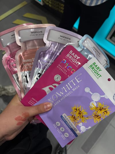 In the city of Nong Pa Khrang, Thailand, a close-up shot captures a hand holding a collection of Watsons "Love My Glow" sheet masks. The masks, varying in color and design, are fanned out, with their upper portions showing brand logos and product names like "Bouncy Serum Mask," "Smoothing Serum Mask," and "Baby Bright Pico." The masks are layered, with the pink and orange ones partially obscured by purple and pink packaging. The hand, adorned with nail polish, displays what appears to be makeup swatches on the palm. In the background, out-of-focus elements suggest an indoor retail environment with yellow and grey floor markings and blue lighting.