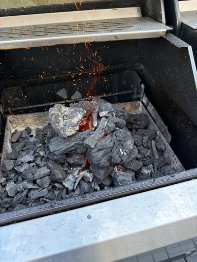 This media file depicts the interior of a charcoal grill, with a pile of glowing charcoal at its center. The charcoal is predominantly black, with some pieces showing bright red embers, indicating they are actively burning. Small orange sparks are visible, particularly to the left of the charcoal pile, suggesting a current ignition or active burning process.

The grill itself appears to be made of metal, with a dark, possibly enameled interior. To the left, a portion of a ribbed metal component, likely a grate or heat shield, is visible, showing some signs of wear and tear, including rust spots. To the right, a wider, lighter colored metal surface forms the outer edge of the grill.

The scene is likely set outdoors, given the natural lighting and the context of a grill. The time of day is not explicitly indicated, but the glowing embers suggest it could be evening or a shaded area during the day. No people are visible in the image. The overall impression is one of preparation for or the beginning of a grilling event. There is no visible text in the image.