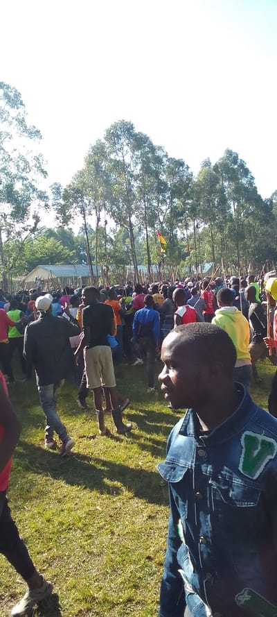 The image shows a large gathering of people in a grassy field. There are many trees in the background, and a single yellow, red, and green flag is visible. The people are standing in a large group, and some are holding long wooden sticks. The individual in the foreground is wearing a denim jacket with a green patch and is facing the camera. The individual in the foreground is facing to the right. The location is in a grassy field in Isukha ICHINA, Kenya, near a village that is visible in the background. The sky is clear and blue. The image was likely taken during the day.