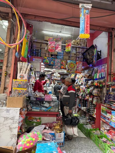 This image depicts the interior of a densely packed retail shop, likely specializing in toys, party supplies, and general merchandise, situated within a market area in Jaipur, India.

The shop entrance is defined by a faded pink facade, with numerous items, including yellow and pink hula hoops and inflatable toys, suspended from the ceiling and doorway. Overhead, string lights illuminate the space. Two security cameras are visible near the top left of the entrance.

Inside, the shop is narrow and rectangular, with merchandise stacked from floor to ceiling on shelves and displayed on counters. A man in a red long-sleeved shirt, likely a shopkeeper, stands behind a glass-top counter on the left, gesturing with his right hand as he interacts with a male customer wearing a patterned shirt. Another customer, an older man in a grey sweater with red shoulders, stands to the right, holding two black motorcycle helmets in his left hand and a white bag in his right. Other male figures are partially visible deeper within the shop.

The shop's shelves are overflowing with packaged toys, games, and novelty items, including doll accessories, plastic kitchen sets, colorful plastic balls, and various other children's products. Large cardboard boxes and plastic crates filled with goods are stacked on the floor. Notable items include a transparent bag filled with numerous neon pink, yellow, and green balls on the lower left, and several green plastic crates filled with merchandise on the lower right. A television monitor is mounted on the right wall, angled downwards. The floor is covered with a light-colored speckled or tiled surface.