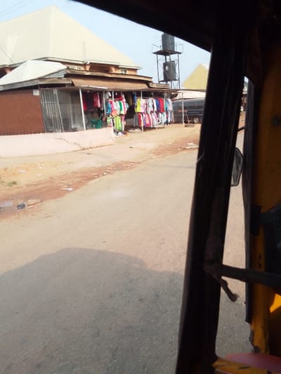 This image presents a street view in Nkwelle Ezunaka, Nigeria, taken from inside a vehicle, likely a tricycle, indicated by the dark frame and yellow interior component on the right. The scene centers on a paved road with minor debris along its left edge. On the left, an open-front shop operates, displaying numerous colorful garments, including t-shirts, blouses, and dresses, hanging on racks. A figure wearing a light green garment is partially visible within the shop. Behind the shop, a multi-story building with a light-colored, sloping roof is prominent. Further down the street on the left, a tall metal structure supports two black water tanks against a clear sky. A dark-colored minivan or bus is parked on the right side of the street, facing away from the viewer. A vehicle's shadow is cast diagonally across the paved road surface in the foreground.