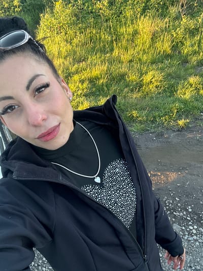 A medium shot captures a woman from a slightly high, angled perspective. She is positioned on a path, with green grass visible to her left and a gray, gravelly surface to her right. A narrow, muddy strip runs parallel to the grass.
The woman wears a black jacket, which is partially zipped up, revealing a black top with a white polka dot pattern. A silver chain necklace with a heart-shaped pendant is also visible. She has dark hair and eyelashes, and is wearing lip color. Sunglasses are placed on her head. She is gazing towards the camera. Her right hand with a ring is visible in the background. The location is not identifiable as Liverpool, United Kingdom.