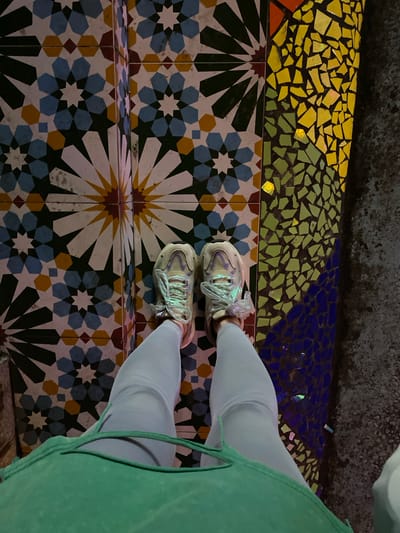 In the city of Baqiao, China, an individual stands on a mosaic-tiled surface. The tiles are arranged in geometric patterns, featuring intricate starburst designs with colors like blue, white, yellow, and dark green. Adjacent to this section, the surface transitions to a mosaic of smaller, irregularly shaped pieces in vibrant yellow, green, and blue. The individual is wearing light blue leggings and a mint green top, with a pair of light-colored sneakers with reflective laces. The perspective is from above, looking down at the person's legs and feet. The lighting suggests an indoor or dimly lit outdoor environment, with some of the mosaic pieces reflecting light.