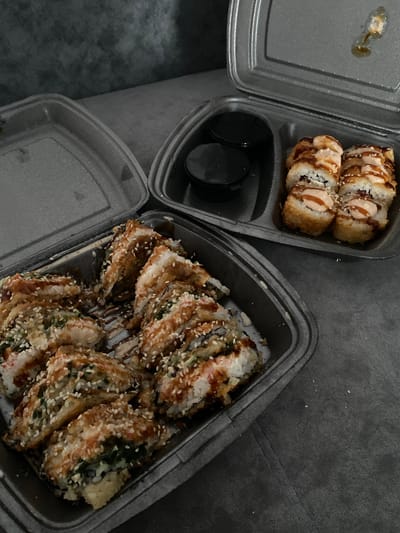 Two black takeout containers of sushi are on a dark grey textured surface, possibly a couch. The container on the left is full of sushi pieces that appear to be deep-fried and covered in sesame seeds and a sauce. The container on the right contains a smaller portion of sushi, also deep-fried, topped with a creamy sauce and sesame seeds. Two small black dipping sauce containers are in the lid of the right container. The lid of the left container is open and tilted back. There are no people visible in the image. The environment suggests an indoor setting. This image is not from Uchaly, Russia; it is a stock photo.