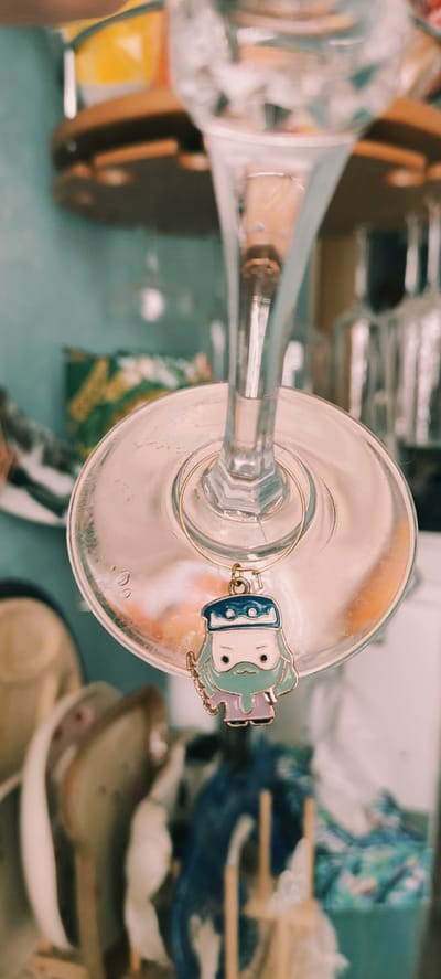 An overhead shot depicts a clear glass object resembling a champagne flute, viewed from above, and lying horizontally. A metallic ring with a blue and mint-green enamel charm of a stylized child-like figure hangs from the rim of the flute. The flute sits on a surface with a teal or pale blue cast. The background features a cluttered assortment of items, including what appear to be wooden trays, a blue bowl or plate, fabric, and boxes, all in a slightly out-of-focus state.