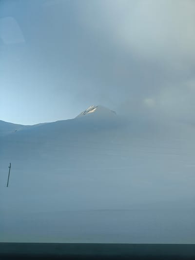 The image captures a vast, snow-covered mountainous landscape in Ganisi, Georgia. The scene is dominated by a blanket of white snow, covering rolling hills and mountain slopes that extend into the distance. A faint, distant mountain peak is visible towards the center-right, its uppermost section catching soft sunlight, suggesting either early morning or late afternoon.
The sky is a pale, hazy blue, transitioning into a lighter, cloud-filled expanse that blends seamlessly with the snow-covered terrain, particularly in the mid-ground, where atmospheric fog or low clouds obscure parts of the view. In the left foreground, a tall, dark utility pole stands upright, emerging from the deep snow. Multiple dark power lines stretch horizontally across the middle of the frame, disappearing into the distant haze.
The overall impression is one of serene isolation and immense scale, characterized by the cold, white expanse. No people or visible text are present. The dark band at the very bottom of the image suggests the photograph was taken from inside a vehicle.