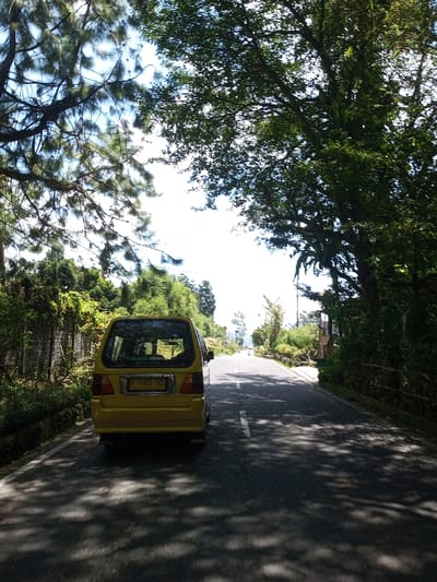 The image displays a yellow minivan parked on the left side of a paved asphalt road, viewed from the rear. The vehicle's license plate reads "F 1963 WX." The road features a broken white line on its left edge and extends into the distance, where it gently curves right. Lush green foliage from various trees, including coniferous species on the left and broadleaf trees on the right, creates a dense canopy over and alongside the road. Dappled sunlight casts numerous shadows across the road surface. On the left, a wall or embankment is covered in climbing vegetation. On the right, further down the road, a utility pole is partially visible amidst the greenery. The sky above is bright and clear. No individuals are present in the immediate foreground or midground. This scene is located in Bagochen Boolingpang Ueling, Bhutan.