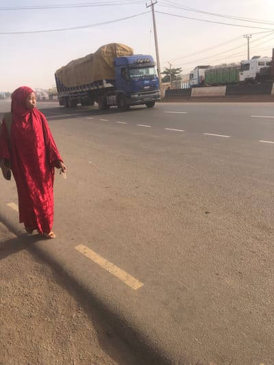 The image depicts an outdoor scene on a paved road in Hanwa, Nigeria, under a hazy, light-colored sky. In the foreground on the left, an individual, appearing female, walks on an unpaved, reddish-brown dirt shoulder. She is dressed in a long, red, patterned garment and a matching red hijab covering her head and neck. Light-colored sandals are visible on her feet. A dark bag is carried on her left shoulder, and she holds a small, light-colored object in her right hand. Her gaze is directed towards the right side of the frame.
Adjacent to the unpaved shoulder is a raised curb with a yellow dashed line, bordering a multi-lane asphalt road marked with white dashed lane lines. A blue Iveco commercial truck, consisting of a tractor unit pulling a trailer covered with a light brown tarpaulin over a significant load, is observed moving from right to left on the paved road in the middle ground. In the far background on the right, several other commercial trucks, including white and green models, are stationary or parked on an adjacent paved area. A utility pole with power lines is visible on the right side of the frame, extending upwards above the road. Distant, indistinct structures are faintly visible through the atmospheric haze.
