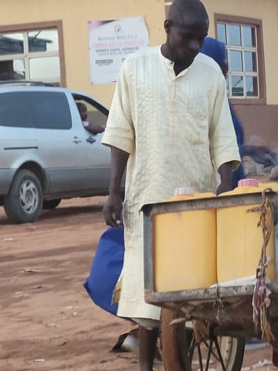 A man in Suleja, Nigeria, is riding a bicycle carrying containers of yellow liquid. The man, who appears to be of African descent, is wearing a light-colored patterned shirt and blue trousers. The containers are loaded in a metal rack attached to the bicycle. In the background, there is a white van parked in front of a building with windows and a poster that reads "Worship With Us, LIVING FAITH CHURCH, WINNER'S CHAPEL, RAFINSEYIN." The ground is reddish-brown, suggesting a dirt or paved surface.