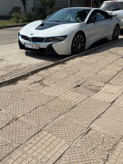 A white BMW i8 coupe (license plate CB 9136 OB) is parked on a textured, light grey paved surface next to an asphalt road. The paving consists of multiple rectangular concrete tiles, predominantly featuring a raised diamond pattern, with some sections exhibiting cracking, erosion, and small weeds growing through the crevices. A concrete curb separates the paved area from the darker asphalt street. To the immediate right and slightly behind the BMW, the rear portion of a silver SUV, possibly a Toyota, is visible. In the background, behind the BMW, there is a weathered white building wall with a dark roofline, green foliage from bushes or small trees, and a dark metal fence. The scene is illuminated by strong sunlight, casting distinct shadows. No individuals are present in the image.

Regarding the specified location, the license plate "CB" indicates that the vehicle is registered in Sofia, Bulgaria, which contradicts the provided information stating the location as Saedinenie, Bulgaria. Therefore, the precise location within Saedinenie cannot be confirmed from the available visual evidence, and the license plate suggests a registration in Sofia.