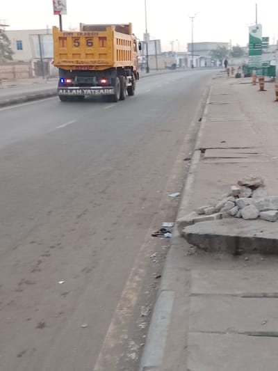 A yellow dump truck with the text "ALLAH YATSARE" on its rear drives along a paved road. Debris litters the roadside. Broken concrete and a pile of rocks rest on the edge of the sidewalk. A building and distant figures are visible further down the road. Construction barrels line the right side of the road. The sky is overcast and hazy.