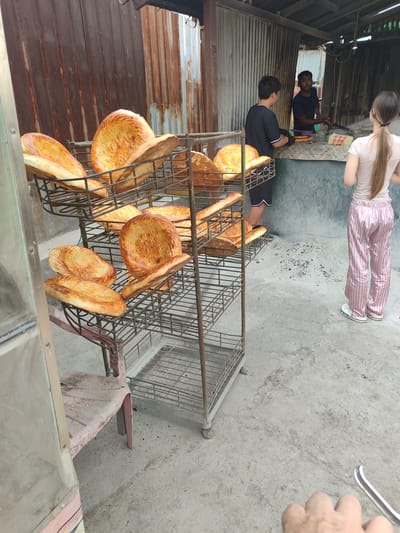 In Tokmok City, Kyrgyzstan, a street-level outdoor baker displays a rack of freshly baked, flatbreads. The breads are golden brown and appear to be of the lavash variety. They are arranged on a tiered metal rack, with some pieces still warm and stacked atop others. In the background, two individuals are seen interacting with a baker, who is preparing dough. The environment is rustic, with corrugated metal siding forming the backdrop of the establishment, suggesting a simple, open-air bakery. The ground is a concrete surface, marked with occasional debris.