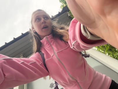 A young person with blonde hair is looking down at the camera. They are wearing a pink zip-up jacket with white drawstring. A white smartwatch is visible on their left wrist. The background includes a white exterior wall and green foliage suggesting an outdoor setting. The location is Bjeliši, Montenegro.