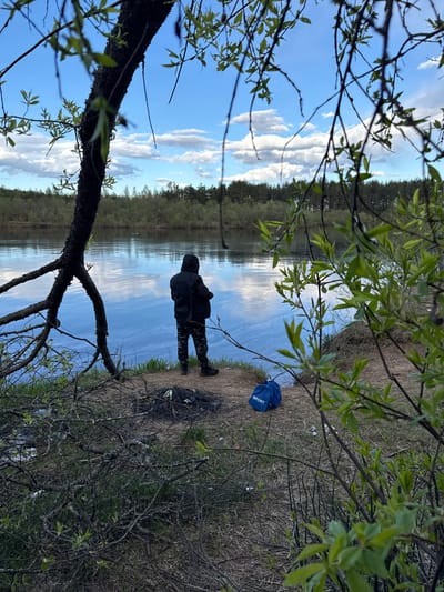 An outdoor scene, possibly in the vicinity of Малая Рукавицкая, Russia, depicts a body of water reflecting a partly cloudy sky. A person stands on the bank, silhouetted in dark clothing. A blue "SPORT" bag is visible near the edge. The scene is framed by branches and foliage.