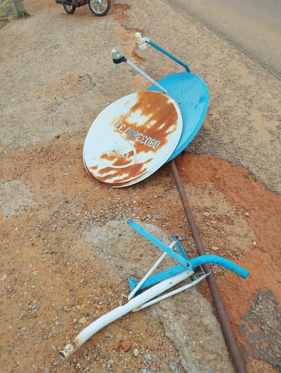 Two satellite dishes, one white and rusted, the other blue, are placed on a dirt and gravel surface next to a paved road. The white dish has faded blue lettering that reads "DSLV so much more" and a serial number. Both dishes are mounted on metal stands, one of which is blue and appears to be broken. A rusty metal pole runs vertically in the background. In the upper left corner, a portion of a motorcycle is visible. The ground is covered with small stones and reddish-brown soil. The scene suggests a discarded or abandoned setup in an outdoor environment.