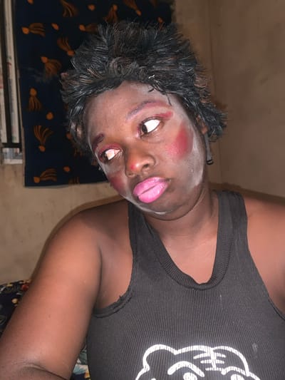 A close-up photograph shows a dark-skinned person with short, dark, wet-looking hair lying on a surface with a dark blue patterned cloth. The person is wearing heavy, unnatural makeup, including bright pink lipstick, exaggerated blush, and pale face powder. They are wearing a black ribbed tank top with a white graphic. The background is dimly lit with a plain wall visible. The person appears to be indoors. There is no indication of the location being Agban, Nigeria or the surrounding area.