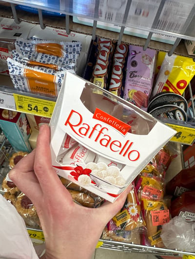 In a supermarket aisle in Novoselye, Russia, a person's hand holds a white box of RaffaELLO confectioneries, which are round coconut-covered chocolates. The box is partially open, revealing several chocolates inside. The background of the image shows shelves stocked with various food items, including packaged wafers with Dutch-style patterns, candy bars, and bottles of ketchup. Price tags are visible on some of the products. The lighting suggests an indoor, commercial environment.