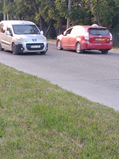 The image shows two vehicles on a paved road with a grassy verge to the left. The vehicle on the left is a silver van with a license plate reading "RWW 308". The vehicle on the right is a red hatchback with a yellow license plate. The scene appears to be in a suburban or semi-rural area, possibly on the outskirts of the city of Bălți, Moldova, given the presence of trees and less dense urban development. The lighting suggests it is daytime, possibly late afternoon or early evening. The overall impression is a candid shot of everyday traffic.