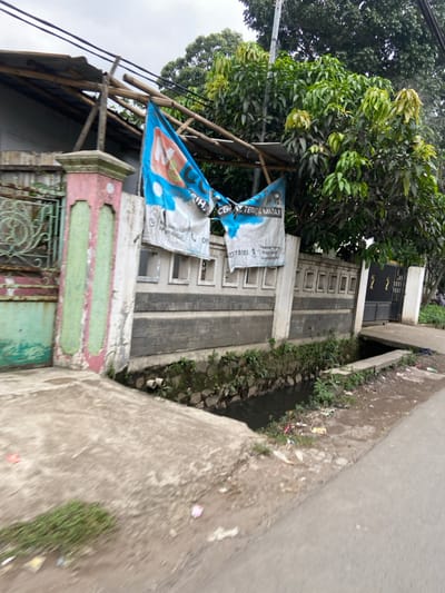 The image shows a street in Margaasih, Indonesia (latitude: -6.962, longitude: 107.548). A concrete drainage ditch runs parallel to the street, with a concrete sidewalk to the right. The ditch is filled with murky water and contains some trash. A large tree with green leaves grows on the left side of the image, casting shadows on the side of the road and the drainage ditch. A concrete wall with a gate in the middle separates the road from a property on the left. The gate appears to be closed. Two large blue and white signs, presumably for a local business, are attached to a wooden frame above the wall. There are no visible people in the image. The sky is overcast. The overall scene is typical of a residential street in a developing country.