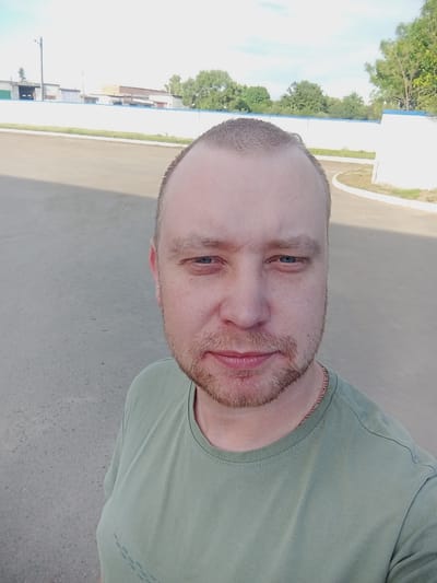 The media file is a selfie of a man, likely taken outdoors during the daytime. The man is positioned in the foreground, facing the camera. He has light skin, a short beard, and light-colored hair. He is wearing a light green t-shirt and a thin chain necklace. The background is blurred but appears to be an outdoor setting with a paved surface and some vegetation. In the distance, there are buildings and trees, suggesting an urban or semi-urban environment. The lighting indicates it is daytime, possibly sunny given the bright sky. The man's expression is neutral. No text is visible in the image. The location context is Mahilyow, Belarus.