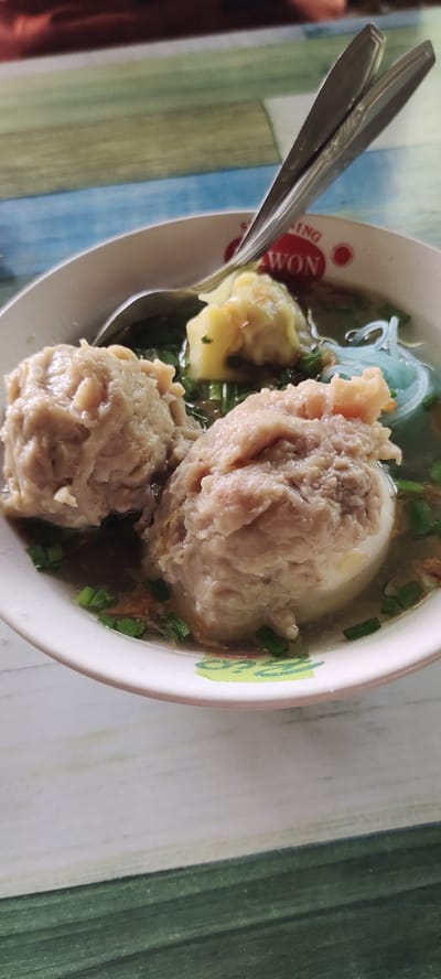 The image shows a bowl of bakso, a popular Indonesian meatball soup. There are two large meatball, one smaller meatball, and a piece of yellow tofu in the bowl. The soup is clear with a light brown hue and is garnished with thinly sliced green onions. The bowl sits on a light blue and green checkered tablecloth. The fork and spoon laying beside the bowl are out of focus. The location of the image, according to the provided coordinates, is in the Lawang District of Malang Regency, East Java, Indonesia. The scene appears to be a food stall or restaurant. The image is likely taken by a customer, as the bowl is centered in the frame, implying the customer is directly looking at the bowl.