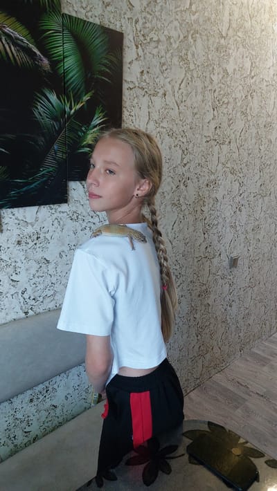 A young girl with blonde hair in a braid is lying down, with a leopard gecko on her back. She is wearing a white t-shirt and black shorts with red stripes. The setting appears to be indoors, with a textured wall and a floor that looks like wood planks. There are two framed pictures of palm leaves on the wall to the girl's left. A phone is visible on the floor to the right. The image is a still shot, and no specific activity or event is taking place, other than the interaction between the girl and the gecko. The lighting suggests it is daytime. The overall mood is calm and relaxed. No visible text is present in the image.