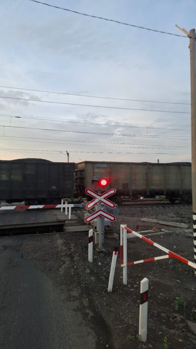 The image shows a railway crossing in Sheragul, Russia, during a cloudy sunset. A train, composed of dark freight cars, is actively passing through the crossing, indicated by motion blur on the cars. The railway crossing is equipped with a red flashing light on a pole, a white and red striped barrier arm that is partially lowered, and a red and white St. Andrew's cross sign. The ground around the crossing is covered with ballast and asphalt. Several white reflective bollards with red markings are positioned near the barrier. Overhead power lines are visible against the twilight sky, which is painted with hues of blue and grey.