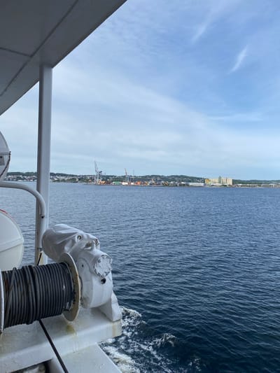 The image is a view from the deck of a ship, looking out towards a coastal city in Stavern, Norway. In the foreground, a white winch with a large spool of dark rope is visible on the ship's deck. The sea, a deep blue with moderate waves, stretches out to the horizon. Across the water, the city of Stavern is visible, characterized by a line of buildings along the coastline, a hill covered with houses behind them, and several industrial structures including large cranes and silos. The sky is a pale blue with scattered white clouds. The overall scene depicts a commercial port area.