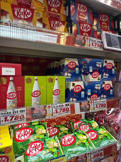 This media file showcases a vibrant display of various Kit Kat chocolate bar flavors, primarily Japanese varieties, presented in a refrigerated retail setting. The arrangement suggests a store specializing in confectionery or souvenirs, likely in Shinjuku, Japan, given the text.

The products are meticulously organized in boxes and individual packs, some stacked high and others laid out horizontally on shelves. Prominent flavors visible include "Strawberry Cheesecake," "Amaou Strawberry," "Mt. Fuji," "Matcha," "Plum Wine," and "Sake Masuizumi." Many of these packages feature distinctive Japanese imagery, such as Mount Fuji, traditional patterns, and illustrations of the featured ingredients.

Several boxes have price tags and product information labels. For instance, a "Strawberry Cheesecake" Kit Kat is priced at ¥1,780 (tax included ¥1,923), and "Matcha" is priced at ¥398 (tax included ¥430). Text in Japanese, along with English translations like "Hot Product" and "Japan. Tax-free Shop," indicates these are intended for both local and tourist consumers.

The lighting suggests an indoor environment, possibly a brightly lit convenience store or a duty-free shop within an airport or train station. The overall impression is one of abundance and variety, highlighting the diverse and often unique Kit Kat offerings available in Japan. The clean presentation and clear labeling suggest a well-managed retail display. There are no people visible in the frame.