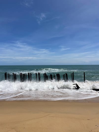 The image depicts a coastal scene in บ้านท่าฉัตรไชย, Thailand, under a clear blue sky with sparse, wispy white clouds. The horizon line separates the sky from the expansive, deep teal-colored ocean. In the mid-ground, a series of dark, weathered vertical posts, varying in height and spacing, are partially submerged in the ocean, extending horizontally across the frame. White, frothy waves are actively crashing against these posts and breaking on the shore, generating significant spray and foam. To the far right, a dark, gnarled piece of wood or debris is partially submerged amidst the breaking waves. The foreground consists of light brown, wet sand, showing ripples and the distinct line of the receding tide. No individuals or discernible wildlife are present.