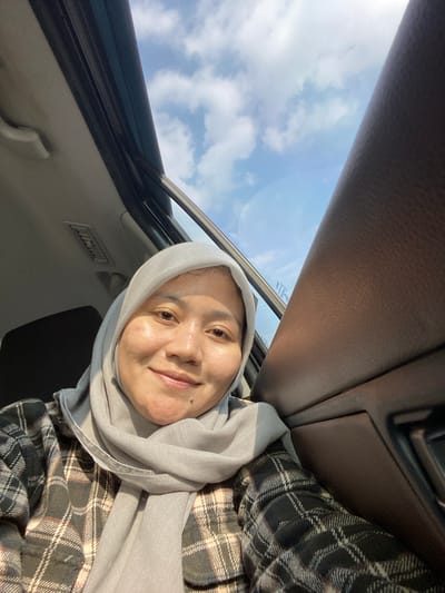 A selfie of a woman wearing a light grey hijab and a plaid shirt is taken inside a vehicle. She is looking towards the camera with a slight smile. The vehicle's sunroof is open, revealing a blue sky with scattered white clouds. The interior of the car is visible, including a brown dashboard and part of a dark-colored seat. The lighting suggests it is daytime. The woman's face is illuminated by sunlight, highlighting her features. The location is identified as Mandar Sari, Indonesia, though no specific landmarks or urban elements are visible in the image to confirm this.