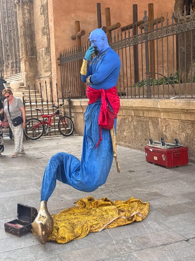 In Málaga, Spain, a street performer dressed as a genie entertains passersby. The performer is clad in a vibrant blue costume, complete with a red sash and golden armbands. Their head is also covered in blue, with what appears to be a painted blue face.

The genie is posed in a dramatic, contorted position, seemingly emerging from or interacting with a large, ornate golden cloth draped on the ground. Next to the cloth is an open, dark-colored chest, and a golden lamp-like object is positioned near the genie's extended arm. A golden staff is also visible, held by the genie.

In the background, a red suitcase with wheels rests against a stone building. A black metal fence, possibly part of a historical building or garden, is also visible, with some wooden posts in front of it. A red bicycle is propped against the fence.

A woman, dressed casually in a grey t-shirt and patterned trousers, stands in the foreground, observing the performance. She wears sunglasses and a dark crossbody bag. The scene appears to be taking place outdoors on a paved walkway, likely during daylight hours, given the shadows. The overall atmosphere suggests a lively street performance in a historical urban setting.