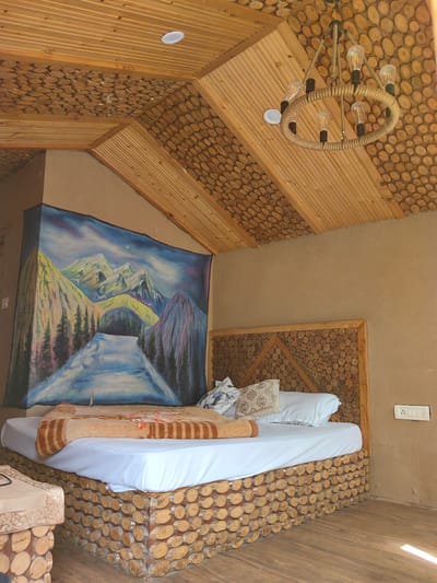 The image depicts the interior of a bedroom in Chhyal, India, featuring a rustic and natural design aesthetic. The room is dominated by wood elements, with the ceiling and parts of the walls clad in wooden planks and circular wood slices. A large, colorful landscape painting of mountains and a river hangs prominently on one wall. The central focus is a bed with a white duvet, a patterned blanket, and several pillows. The bed frame and headboard are constructed from wood slices, echoing the decorative elements of the ceiling. A rope-and-bulb chandelier hangs from the ceiling, adding to the rustic ambiance. A bedside table, also made of wood slices, is partially visible on the left. A light switch is mounted on the right-hand wall. The flooring appears to be wooden planks. The overall impression is one of a cozy, nature-inspired retreat.