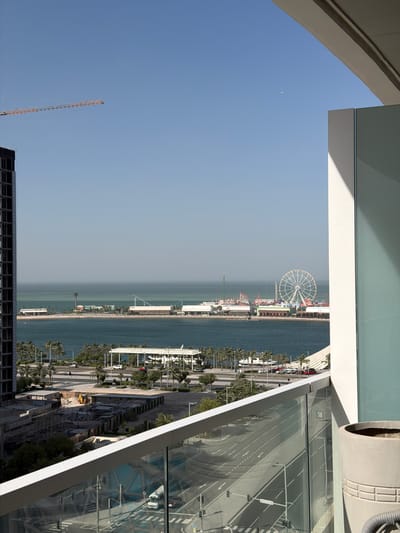 This image captures a wide-angle view of a coastal city from a high vantage point, looking out over a body of water. The sky is a bright, clear blue, with a faint crescent moon visible in the upper left.
In the immediate foreground, architectural elements of a modern building are prominent, including white beams and large glass panels that reflect a street scene below, showing palm trees, traffic lights, and road markings. A large, grey cylindrical structure is also partially visible in the upper right.
The midground reveals an expansive waterfront characterized by a serene sea. Along the coastline, a vibrant development stretches across the frame. A large Ferris wheel dominates the center-right, indicating the presence of an amusement park or recreational pier. Numerous palm trees line the thoroughfares and coastal areas. Several multi-lane roads are visible, complete with traffic lights and distinct road markings, and a few cars can be seen.
The cityscape includes a mix of completed buildings and ongoing construction. A large, dark, multi-story building frame under construction occupies the lower left corner, and a tall construction crane extends into the sky from this area. Another construction site with a yellow excavator is visible towards the lower right. The distant horizon over the water appears somewhat hazy. The time of day is daytime, with bright sunlight illuminating the scene. No discernible text is visible. The scene suggests significant urban development and recreational activity in a coastal setting, consistent with Lusail, Qatar.