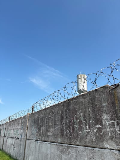 This media file features a concrete wall topped with barbed wire, set against a clear blue sky with a few wispy clouds. The perspective is looking upwards from the base of the wall.
The wall appears to be old and weathered, with patches of discoloration and what looks like graffiti scrawled on its surface. The barbed wire is a prominent feature, forming tangled spirals along the top of the wall, suggesting a security perimeter. Attached to the wall, midway up, is a cylindrical object, possibly a light fixture or sensor, encased in metal.
The bright blue sky indicates it is daytime, likely a sunny day. The scene evokes a sense of confinement or restriction due to the presence of the security wall and barbed wire. The graffiti adds a touch of human presence, but the overall atmosphere is stark and somewhat somber.
While no people are visible, the graffiti on the wall might suggest past human activity. The image is taken at a low angle, emphasizing the height and imposing nature of the wall. The location context provided, Ashmyany, Belarus, gives a geographical reference but doesn't offer specific visual cues within the image itself. There is visible text in the form of graffiti on the wall, but it is indistinct and illegible in this view.
