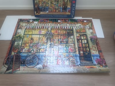 The image depicts a completed jigsaw puzzle featuring a charming bookstore facade. The puzzle is laid out on a light-colored wooden surface, and a portion of its box is visible in the upper portion of the frame. The puzzle itself displays a colorful and detailed scene of "The Greatest Ever Book Shop," complete with bookshelves overflowing with books, a bicycle parked outside, flower baskets, and a welcome mat at the entrance. The storefront is decorated with awnings and banners, creating a welcoming atmosphere. The image does not contain any specific information that would allow for identification of its location in the city of Mississauga, Canada.