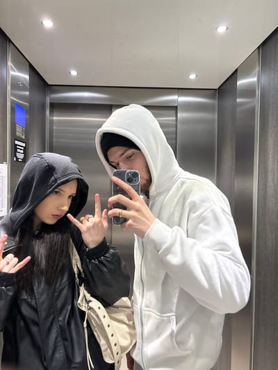 A selfie taken in an elevator mirror shows two people in hooded sweatshirts. The person in the white hoodie holds a phone and wears a ring. The second person wears a black hoodie and jacket with a shoulder bag, also sporting rings and making a hand gesture. The elevator interior includes lighting and a control panel visible in the reflection.