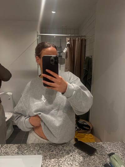 The image shows a woman in a grey sweatshirt and grey sweatpants standing in a bathroom in Skanderborg, Denmark. The woman is holding a black smartphone in her right hand and is looking at the camera. Her left hand is on her stomach. She is standing in front of a bathroom sink with a white countertop and a speckled grey backsplash. Behind the woman is a white toilet, a brown shower curtain, and a green towel. The bathroom has a white tiled floor. There is a second smartphone lying on the counter next to a blue and green bag and a yellow object. The image was taken in a bathroom in Skanderborg, Denmark (latitude: 56.049, longitude: 9.926). The image appears to be taken using a mirror.