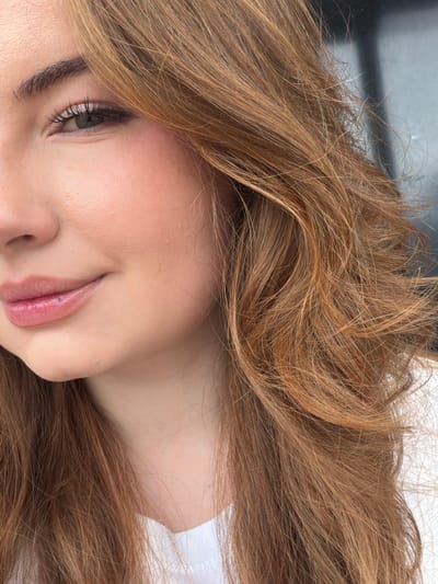 A close-up, waist-up shot shows the right side of a woman's face and chest, her hair cascading down her right shoulder. Her gaze is directed downwards, and her lips are slightly parted, revealing a hint of her teeth. A faint blush dusts her cheeks, and a subtle sheen highlights her lower lip. Her eyebrows are dark and well-defined, framing her eye. The background is blurred, featuring a dark, vertical element that could be part of a building or a large object. The lighting appears to be natural, casting a soft glow on her skin. The image does not contain any information that would allow for the identification of a specific location in Sarpsborg, Norway.