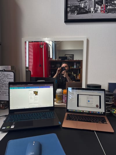 This media file captures an indoor workspace in Katwijk, Netherlands, featuring a desk setup with two laptops and a mirror reflecting a person.

The main subject is a man, visible in the mirror reflection, taking a selfie with a smartphone. He has a beard and a noticeable tattoo on his left arm. The setting is a home office, with a desk against a textured white wall.

On the desk, there is a rose gold/gold MacBook Air and a black Lenovo laptop, both open and displaying documents or web pages. A blue mousepad with a gray "Trust." branded mouse is beside the Lenovo. Other items on the desk include a black circular speaker and several supplement containers, such as "CALM & RELAX" and "NOHYD CREATINE MONOHYD 210g". A framed black and white photograph of a vintage car scene hangs on the wall to the left. The mirror also reflects bookshelves and a large red appliance, possibly a refrigerator, behind the desk. A white board or notebook with handwritten notes is visible in the foreground. The ambient lighting suggests it is daytime.

Visible text includes:
*   **Handwritten on the notebook:** "Ниши.", "ВЫБОР Ниши.", "на торговые марки.", "Аудитории.", "книги и Пеевариин.", "вка кинги IC THEAIKALL", "ПРОВЕРКА не", "BAUUE", "Kindle Create", "Для печатией", "17.02.26", "PRIVAL TATA 3", "04.2026 Seιπια", "Заметки Таймер".
*   **On the MacBook Air screen:** "MacBook Air", "Ресурси" (Resources), and part of a phrase "Подать запрос на ут" (submit a request for ap).
*   **On the Lenovo laptop screen:** "Страница 1 из 4" (Page 1 of 4), "Файл" (File), "Изменить размер" (Change size), "Редактирование" (Editing), and the "Lenovo" brand logo.
*   **On product labels:** "Trust." on the mouse, "CALM & RELAX", "Helps to ma during periods", "SKI", "NOHYD CREATINE MONOHYD 210g" on the supplement containers.