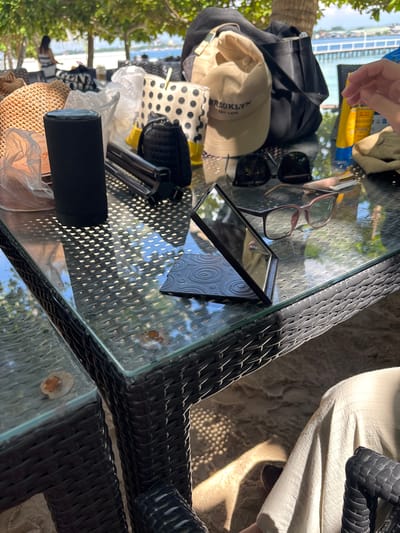 The image shows a collection of items on a glass-topped table situated outdoors, likely at a beach resort in Samal, Philippines. The table has a woven rattan base and is casting dappled shadows from the surrounding foliage and sunlight onto the sandy ground.
Among the objects are two pairs of sunglasses, one with dark lenses and a thin metal frame, and another with thicker, pinkish frames. A small, square compact mirror with a decorative, circular pattern on its case is open on the table. A tan baseball cap with "BROOKLYN" embroidered on it is also visible, alongside a black and white polka dot bag. There's also a black fabric item, possibly a bag or a cover, and a straw hat. A black cylindrical object, possibly a speaker or a thermos, rests on a plastic bag. Two yellow tubes of what appears to be sunscreen are also present. A hand is reaching into the frame from the top left, suggesting someone is interacting with the items.
In the background, a glimpse of a body of water, possibly the sea, and a structure with railings, perhaps a boardwalk or a pier, can be seen. Lush green trees frame the scene, indicating a tropical environment. The lighting suggests it is daytime, with bright sunlight creating strong highlights and shadows, typical of a sunny day. The presence of sunscreen and beachside elements like the hats and sunglasses strongly implies a vacation or leisure activity.