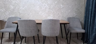 A dining set is positioned in a room. The set includes a light-brown wooden table with dark legs and four light-gray chairs. Two chairs are visible from the front and two from the back. The chairs have a channeled design on the backrest. Behind the dining set, the wall is covered with textured wallpaper featuring a marbled pattern in shades of white and beige. To the right of the dining set, a dark blue velvet curtain hangs. The floor appears to be wood-effect laminate. The image is captured from a slightly low angle, with the table and chairs filling the foreground and midground. The lighting is neutral, suggesting an indoor setting. The scene does not provide any information about the city of Navoiy City, Uzbekistan.