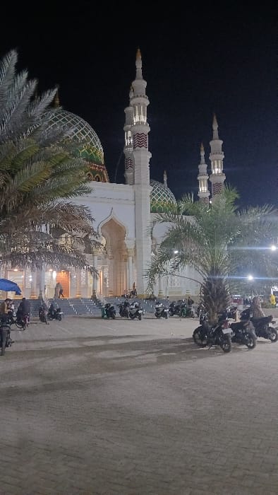 The image displays a large, illuminated mosque at night in the city of RawangItiek, Indonesia. The mosque is predominantly white, featuring multiple tall minarets adorned with white and red horizontal striped patterns, topped with golden finials. Several domes, primarily green with intricate golden patterns, crown the structure. A prominent arched main entrance leads to a wide set of stairs.

The foreground consists of a vast, light-grey paved area, serving as a parking lot. Numerous motorcycles, approximately 20-30, mostly dark-colored, are parked across this area and closer to the mosque steps. Several large palm trees are distributed throughout the scene, with a prominent one in the mid-right and another on the far left, partially obscuring a mosque dome.

Approximately 10-15 individuals are discernible within the scene, primarily congregated near the mosque's entrance steps or moving across the paved area. One individual is visible under a blue canopy-like structure on the far left. The environment is artificially lit, with streetlights and mosque illuminations casting light across the foreground and building, contrasting with the dark night sky.