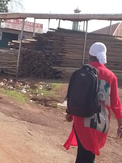 A person wearing a white head covering and a red long-sleeved shirt with a black backpack walks along a dirt road in Dungu, Ghana. Behind the person are stacked piles of lumber and logs, and a sign reading "SOL CEMENT AWUNI ALU ENTERPRISE" is visible on a building. The environment includes vegetation, debris on the ground, and a building with a red roof in the background.