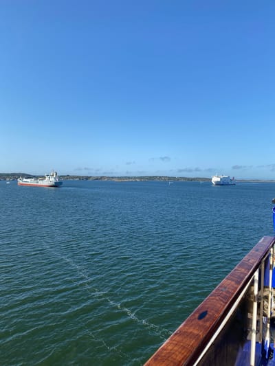 The image shows a body of water, likely the sea, with a clear blue sky above. In the distance, two ships are visible. One is a large cargo ship with a grey hull and a red waterline, moving from left to right. The other is a white passenger ferry, with "Stena Line" written on its side, moving from right to left. The background features a coastline with rocky islands and vegetation, characteristic of the Asperö area of Sweden. Part of a wooden railing is in the foreground, suggesting the image was taken from another vessel. The water is rippled, indicating some movement.