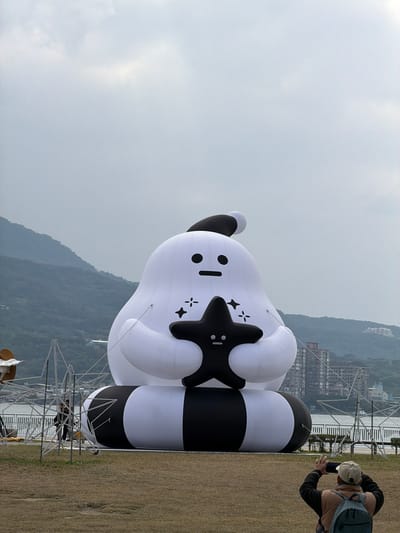 A large, white inflatable character stands on a grassy field near a lake and mountains. The character has black and white striped legs, a black star-shaped patch with a small face on its chest, and a simple facial expression on its head. A person wearing a tan hat and backpack takes a photo of the character with their phone. Metal scaffolding is visible behind the character, and buildings are seen in the distance across the lake. The sky is overcast.